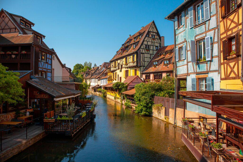 Une vue pittoresque d’un canal bordé de maisons à colombages colorées dans une ville d’Alsace, en France. Le ciel est bleu et ensoleillé, et plusieurs restaurants avec des terrasses donnent sur l’eau, créant une ambiance chaleureuse et authentique.